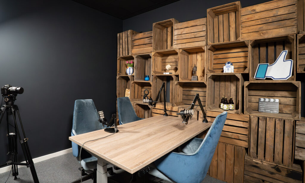 Dienst_podcast Moderne podcaststudio met houten kistenwand, twee blauwe stoelen, microfoons op verstelbare armen en een videocamera op statief.