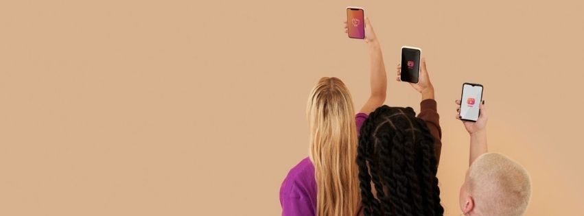 3 mensen die een telefoon omhoog houden en op het scherm social media kanalen open hebben. De 3 mensen kijken naar hun eigen scherm.