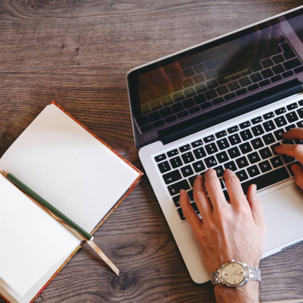 1 Handen die op een laptop typen aan een houten bureau met een open notitieboek, potlood en polshorloge.