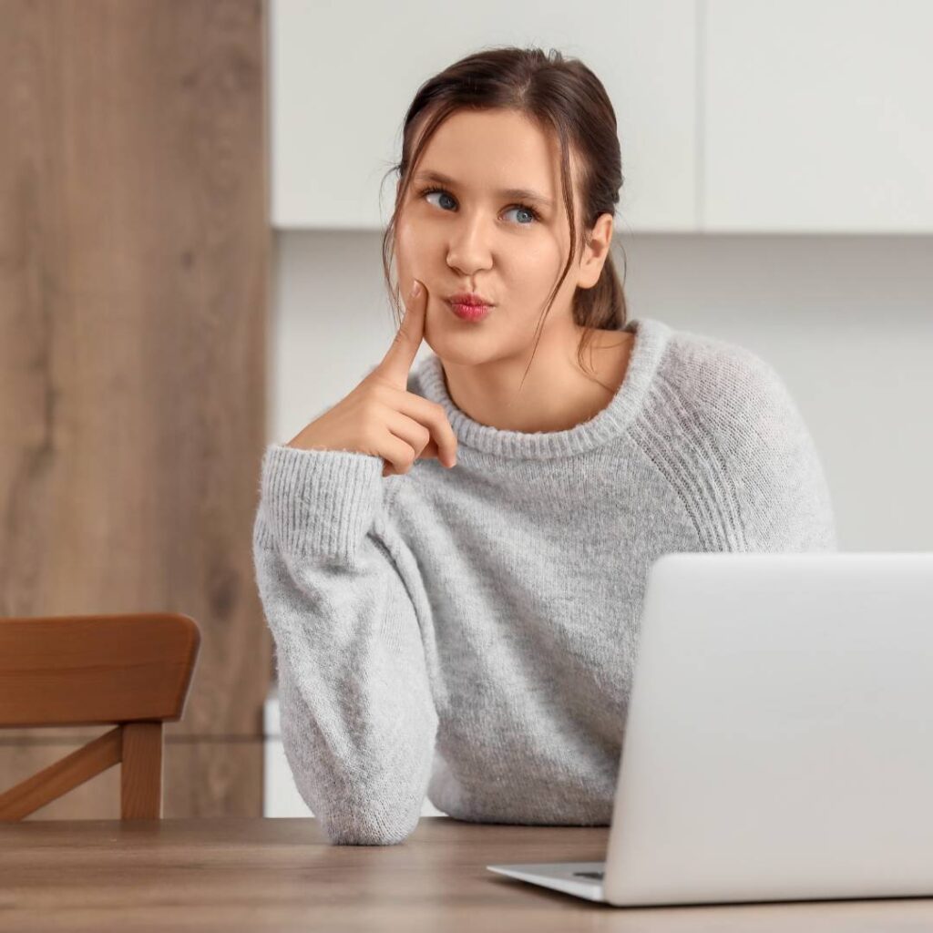 3 Vrouw in grijze trui zit aan tafel met laptop en kijkt nadenkend opzij met vinger tegen haar wang.